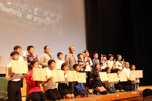 壇上で山﨑江東区長を囲んで記念写真を撮る優秀校の代表者たち。子どもたちが掲げる表彰状は木の紙からできている。
